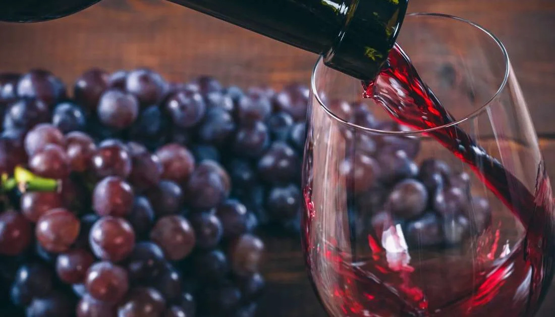 Red wine pours into a glass beside a bunch of ripe grapes on a rustic wood table—capturing the richness, freshness, and elegance of the winemaking process